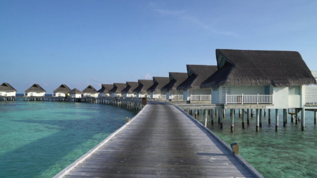 A row of tropical bungalow houses with thatched roofs above clear blue ocean water, 4K Desktop Wallpaper
