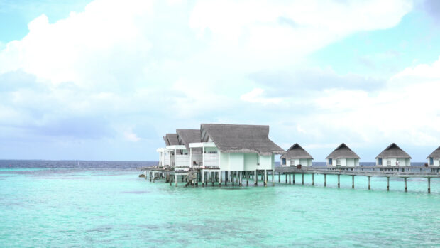 Tropical bungalow houses stand on stilts above clear turquoise ocean water with a cloudy sky, 4K Desktop Wallpaper