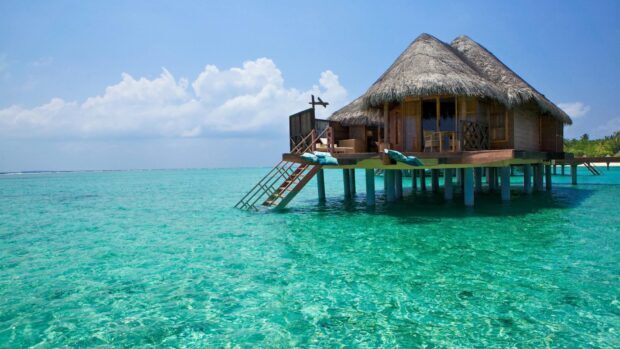 A tropical bungalow surrounded by turquoise ocean water under a blue sky with clouds, HD Desktop Wallpaper
