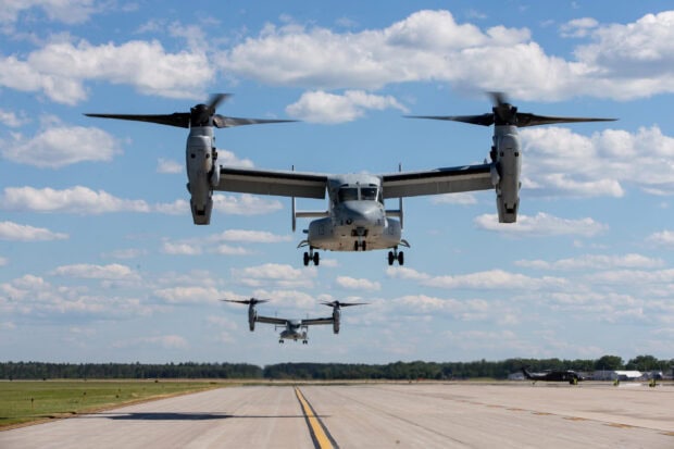 V 22 Osprey aircraft flying low over a runway on a clear day with blue sky and scattered clouds