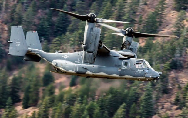 A military aircraft with osprey rotors flying over a forested mountain area