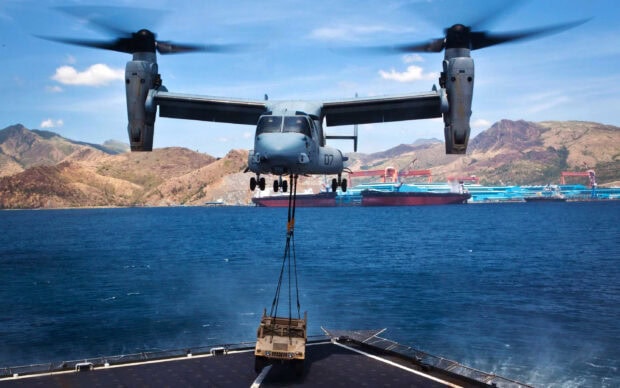 A military V 22 Osprey helicopter lifting a vehicle over the ocean near mountains