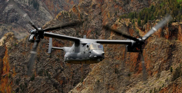 A V 22 Osprey flying closely to a rocky mountain cliff covered with sparse trees