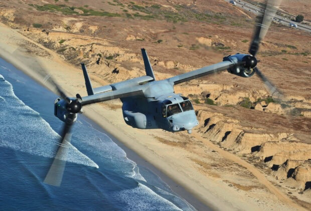A V 22 Osprey aircraft flying low over a coastal beach and rugged cliffs
