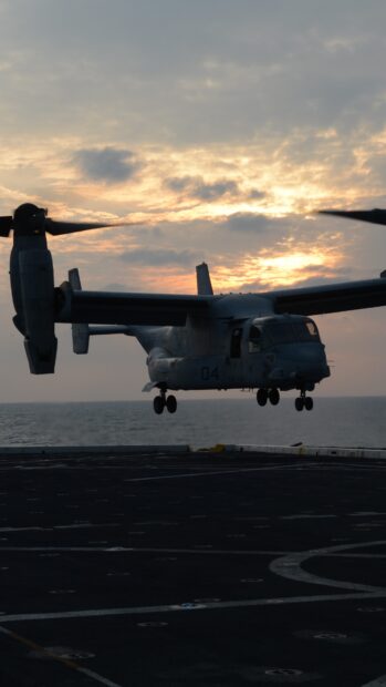 A V 22 Osprey aircraft flying over the ocean at sunset with a cloudy sky