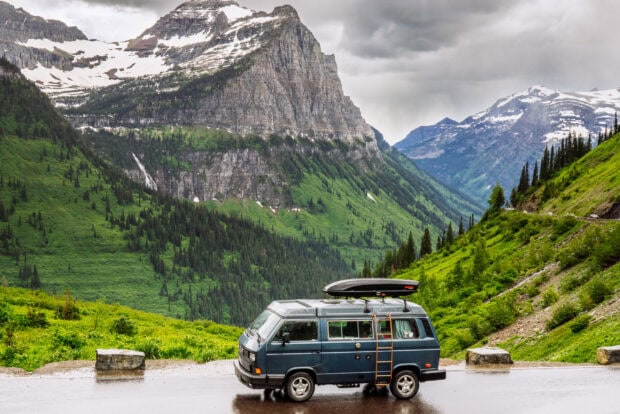A blue van is parked near lush green mountains under cloudy skies in Glacier National Park, 2K Desktop Wallpaper