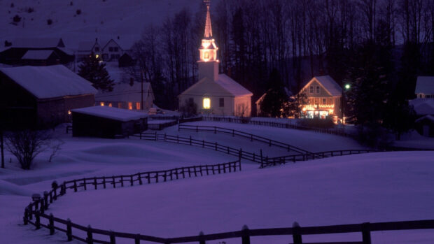 A peaceful Vermont town covered in snow with warmly lit houses and a glowing church steeple at dusk, 2K Desktop Wallpaper