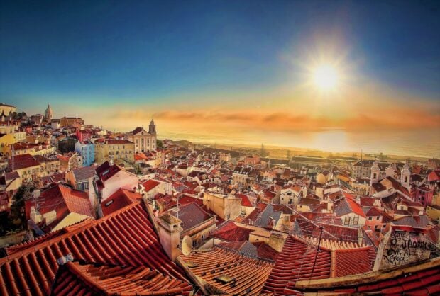 The vibrant cityscape of Lisbon Portugal shows red rooftops under a bright sun near the ocean, 2K Desktop Wallpaper