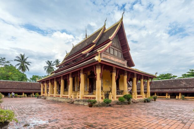 Historic Vientiane temple with ornate roof and tall pillars under cloudy sky