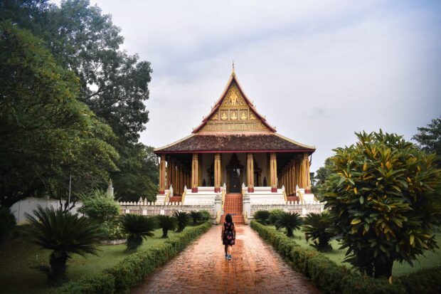 A person walking towards a traditional Vientiane temple surrounded by lush greenery