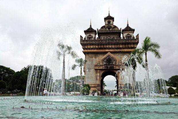 Patuxai monument in Vientiane with fountain and palm trees on a cloudy day