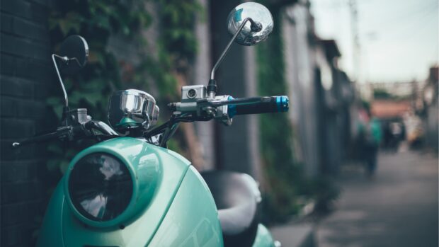 A vintage green scooter is parked on a blurred street beside an old brick wall with plants, 4K Desktop Wallpaper