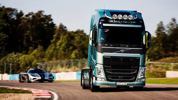 A Volvo truck driving on a race track with a sports car following behind in a forest setting