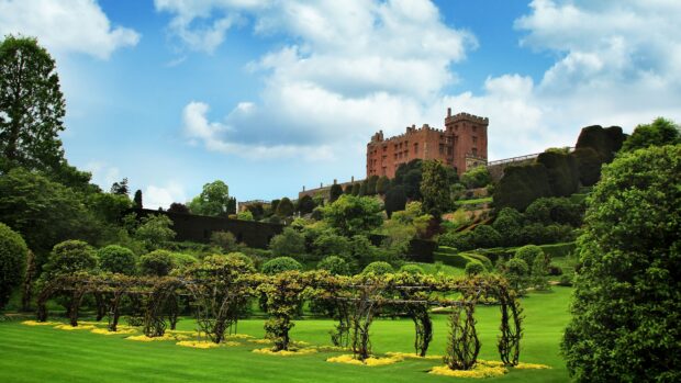 A historic castle in Wales surrounded by vibrant green gardens under a cloudy blue sky, 4K Desktop Wallpaper