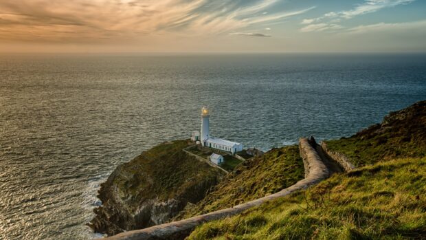 A lighthouse stands on a cliff overlooking the ocean in Wales with sunset skies, 4K Desktop Wallpaper