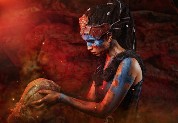 A warrior from Hellblade adorned with tribal paint holds a burlap sack in a fiery setting, 2K Desktop Wallpaper