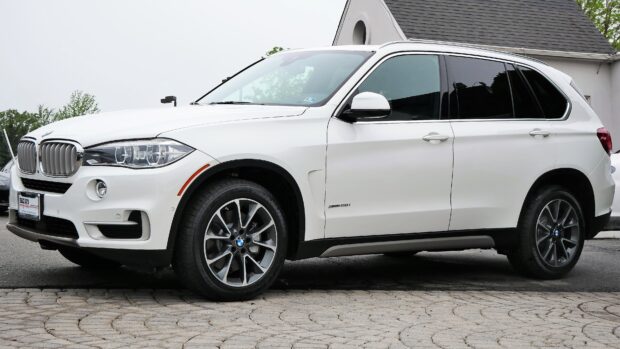 A white BMW X5 SUV is parked on a paved driveway beside a house with trees in the background