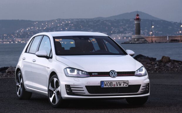 A white Volkswagen Golf GTI is parked on a rocky shore with a lighthouse and city lights in the background, 2K Desktop Wallpaper