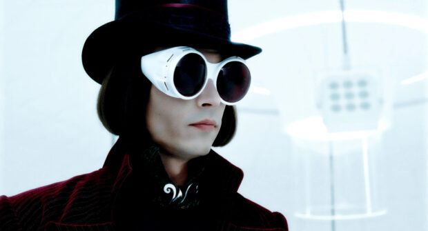 Willy Wonka wearing white round glasses and a black top hat in a bright room