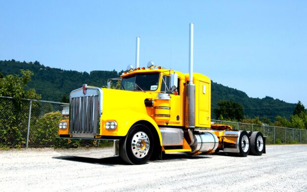 A yellow Kenworth truck is parked on a gravel road with trees and hills in the background, 2K Desktop Wallpaper