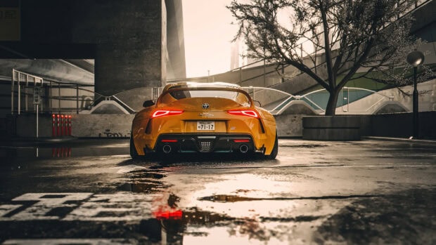 Sports car parked after rain, yellow Toyota Supra sports car in urban setting with reflections on wet pavement, modern city architecture, and sunset lighting.