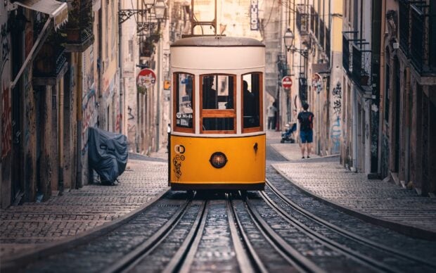 A yellow tram moves through a narrow street with cobblestone tracks in Lisbon Portugal, 2K Desktop Wallpaper