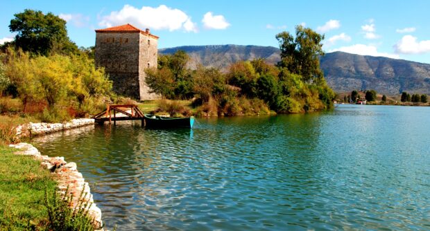 Historic tower and boat near calm river in Albania surrounded by trees and mountains