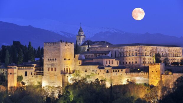 The Alhambra castle lit up at night under the full moon and mountain backdrop