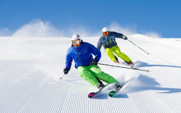 Two skiers wearing helmets and bright gear enjoying alpine skiing on a groomed snowy slope