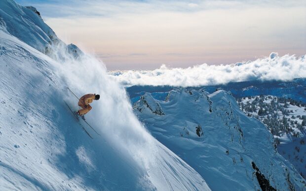 A skier in brown outfit skiing down a steep snowy alpine slope with snow clouds around