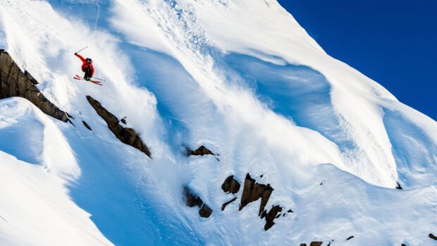 A skier performing a jump on an alpine slope with snow and rocks visible