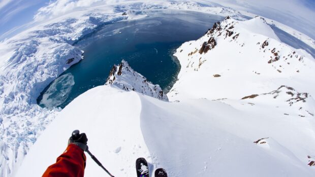 A skier wearing a red jacket skiing down a snowy mountain with a large icy lake in the background