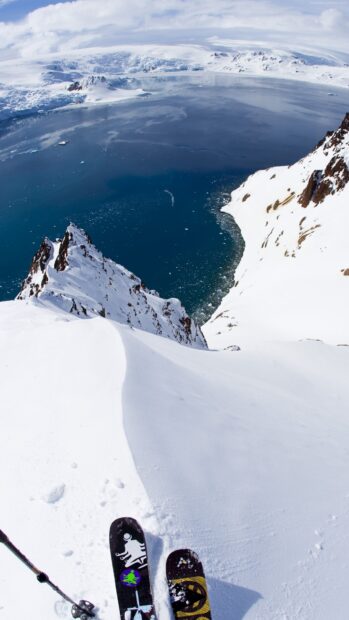Steep alpine skiing slope covered in snow with a view of icy mountain lake and skis in foreground
