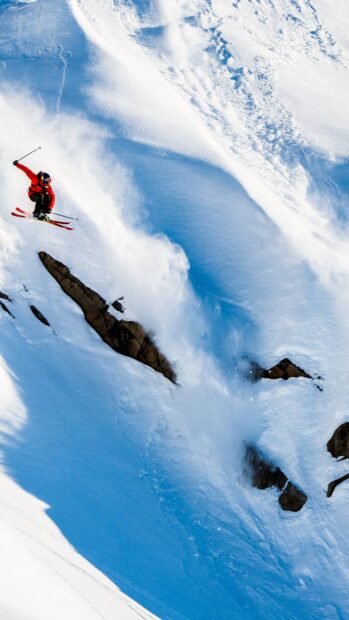 Skier performing a jump on a snowy alpine slope with fresh powder and rocks visible