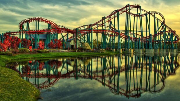 A colorful amusement park roller coaster with red tracks and blue supports reflected on a lake