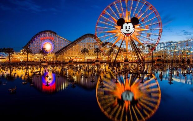 A vibrant amusement park with a large ferris wheel featuring a smiling face reflecting on the water at dusk