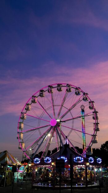A vibrant amusement park with a Ferris wheel lit up in pink lights at sunset