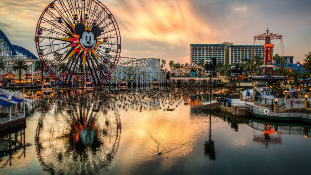 A vibrant amusement park with a large Ferris wheel featuring a cartoon face over a reflective lake at sunset