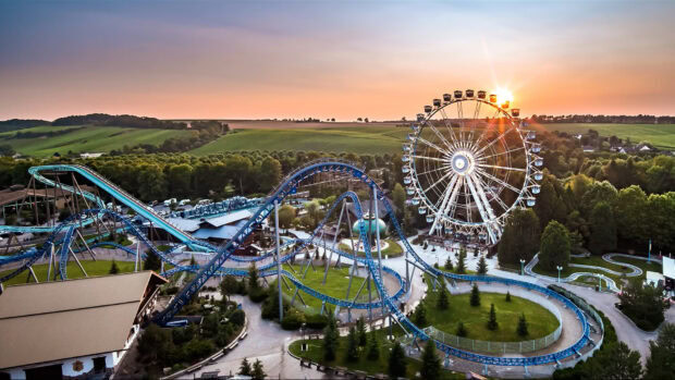A roller coaster and ferris wheel in an amusement park at sunset with lush green surroundings