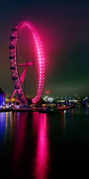 A brightly lit Ferris wheel at night reflecting pink lights on the river surface