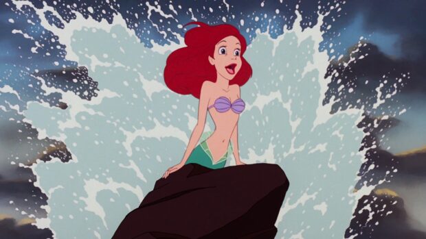 Ariel from The Little Mermaid stands on a rock with water splashing behind her in a classic scene