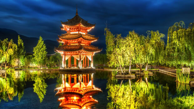 Traditional Asian architecture reflects in pond water during night with illuminated trees and mountains