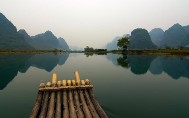 A bamboo raft floating on a calm river surrounded by Asia mountains and trees reflecting on water