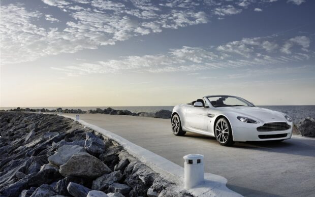 White Aston Martin Vantage convertible car parked on a coastal road with rocky shore and cloudy sky