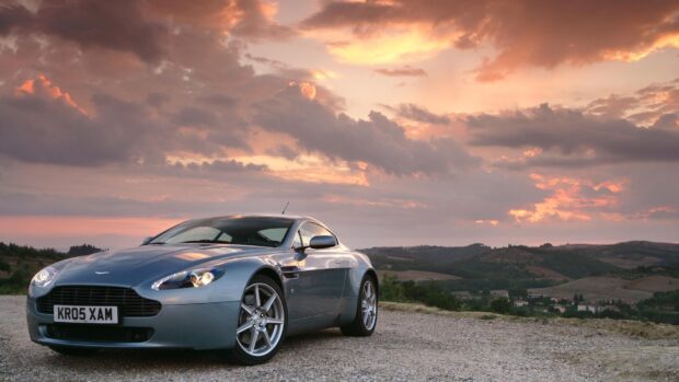 A sleek Aston Martin Vantage parked on a gravel road at sunset with scenic hills in the background