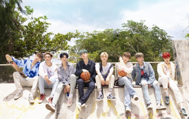 Ateez group members sitting outdoors with basketball and skateboard in a bright urban setting