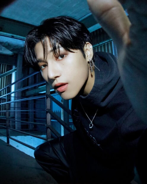 Male Ateez member with black hair wearing earrings and a black hoodie in an industrial setting