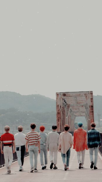 Ateez members walking on a wooden bridge towards a rusty metal structure and mountainous background