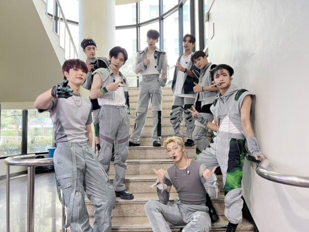 Ateez members posing on a stairway inside a building wearing coordinated gray outfits