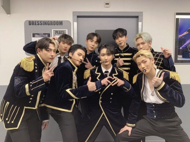 Ateez members posing together in matching navy and gold outfits backstage dressing room